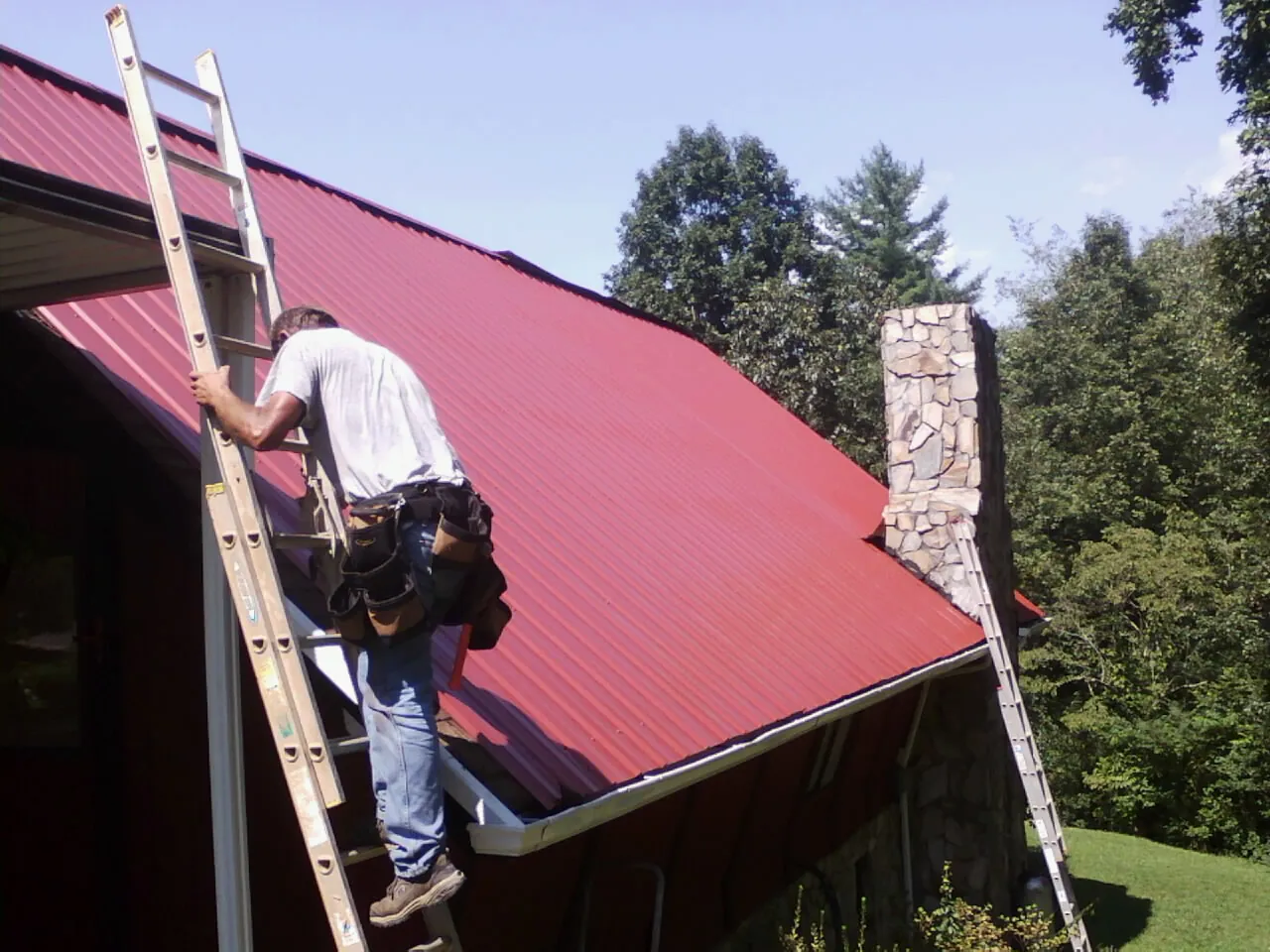 Roofer on a ladder beside a red metal roof and stone chimney — Soffit & Fascia Repair by Roofing Brothers in Central Bridge